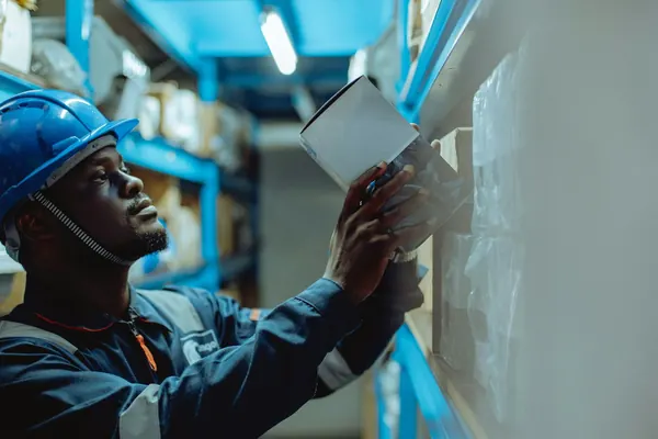 A worker in a blue hard hat checking a list beside warehouse shelves
